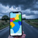 Route de voyage sinueuse sous un ciel chargé d’orage, avec un smartphone au premier plan montrant une carte météorologique abstraite aux couleurs d’alerte.