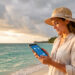 Une voyageuse consulte une tablette face à un littoral tropical, sous un ciel partagé entre soleil et nuages.